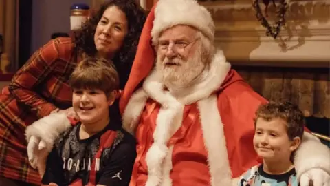 Imagination Alive Productions Father Christmas, in a red suit and hat, poses with a family for a photo.