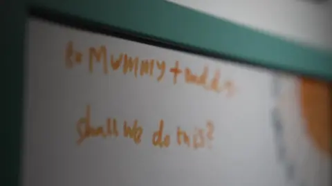 A child's drawing which reads: "To mummy and daddy. Shall we do this?" In the corner of the picture is a drawing of a sun, which is out of focus. It looks to be framed in a teal-coloured frame.