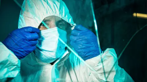 Blast Films Crime scene coordinator, Jo Ward, is dressed in a protective white suit. She is gripping the hood of this suit with both hands and is wearing blue plastic gloves. Jo is looking to the right of the image and staring seriously at a cracked window pane. It is evening and the sky is dark behind her.