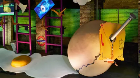 A surrealist art exhibit on a disused Tube platform featuring two giant cardboard eggs, one cracked open with a yellow yolk and the other with a nail sticking out of it. There are oversized ladders, megaphones and large chicken's legs as part of the exhibit. 