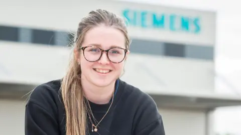 Siemens Mobility Liv has long blonde hair tied back, has wide rimmed, circular-framed glasses. She is smiling for the camera, with a Siemens sign on the side of a building in the background.
