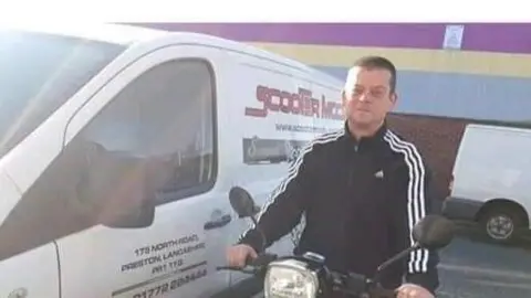 HSE handout Lee Warburton, with short brown hair, wears a black tracksuit top as he sits on a motorbike parked up near a white van near a warehouse.