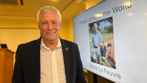 Steve smiles at the camera as he stands next to a projector screen. It has a photo of him and a Jersey cow and the text reads 'Jerseys Around the World'. Steve is wearing a dark navy blue jacket with a crosshatched shirt on. Steve has short white hair.