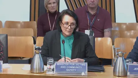 Community safety minister Siobhan Brown - a woman with shoulder-length black hair and glasses, wearing a dark suit and a green blouse, giving evidence in a Scottish Parliament committee room
