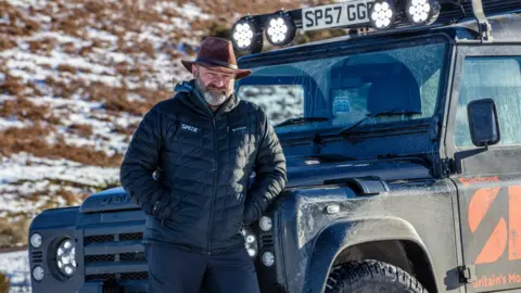 Bearded race director Phil Hayday-Brown in a brown brimmed hat and a puffer jacket stands in front of a Land Rover with lights across its front in a snowy countryside