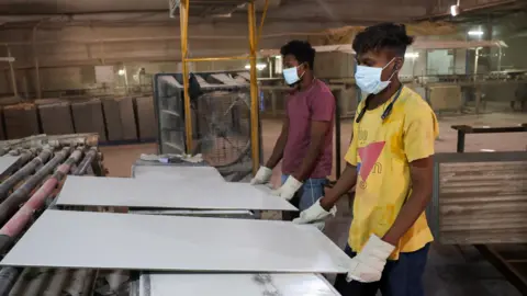 Reuters Two men - one wearing a yellow t-shirt and another wearing a maroon t-shirt - work at a ceramic tile production line at a factory in Gujarat' Morbi on 5 March 2026.