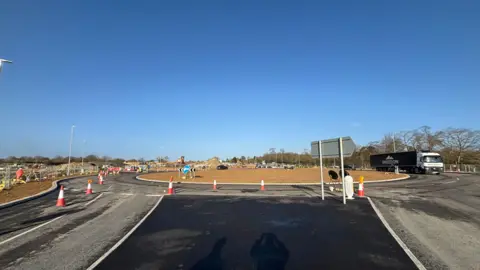Luke Deal/BBC A wide view of a newly constructed roundabout on a sunny day. A lorry can be seen on the right as it moves around the roundabout. Cones block off the outside lane. There are no clouds in the sky.