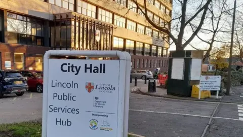 Google Street View A golden sun shines on the glass windows of a large modern council office building. Cars are parked on asphalt outside. In the foreground, a white sign reads: "City Hall, Lincoln public services hub".
