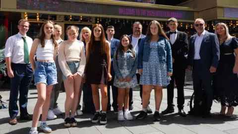 Into Film A group of thirteen adults and young people stood outside the location of the film awards ceremony. The sun is shining on them, and behind them is a sign which says 'Welcome to the 2025 Into Film Awards'.