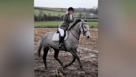 Katie is seen on a grey horse, she is smiling with her thumb up. There are green hills behind her.