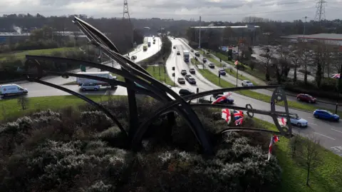 A large metal artwork in the middle of a roundabout. There are flags hanging off part of it. Cars can be seen approaching and going around the roundabout.