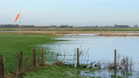 A flooded field with a fence in the foreground and an orange airfield wind sock in the distance 