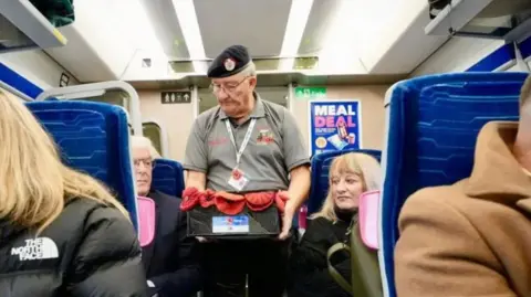 A bespectacled man, wearing an Army beret and a grey T-shirt, is carrying a tray of poppies as he walks down an aisle on a train, watched by seated passengers.