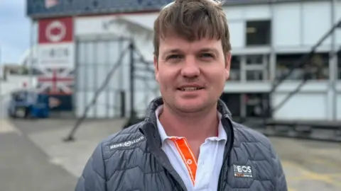 Man wearing a dark grey coat with white shirt with orange ending he has brown hair and sunglasses on top of his head