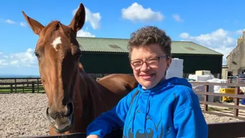 Louise Schilt, who has short, brown, curly hair and glasses is wearing a blue hoodie and looking at the camera while standing next to a brown horse.