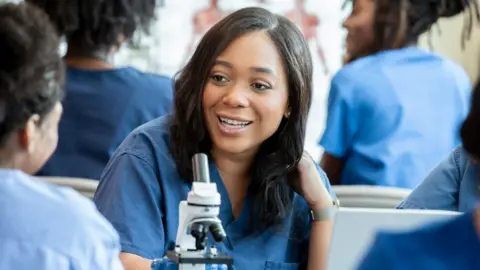 Getty Images A young smiling healthcare worker is leaning on a desk with a microscope on it. She has long, dark brown hair, wears blue overalls and is surrounded by others in the same clothing.