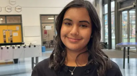 A teenage girl with dark hair and a black top smiles at the camera