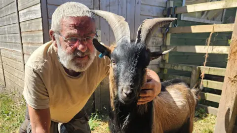 Wayne Eckersley, sitting down, with his arm around a goat looking straight at the camera