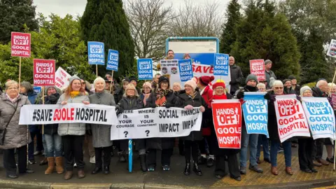 BBC/Anne-Marie Tasker Group of people standing in front of trees holding placards, banners and signs with slogans about saving Goole hospital.