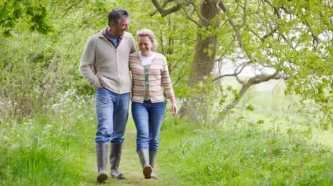 A man with dark hair and a woman with blonde hair both wearing beige jumpers, jeans and wellington boots walking through a grassy area with trees behind them
