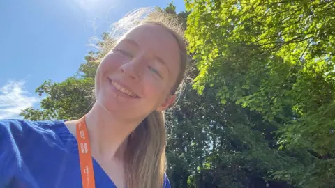Ulster University A smiling young woman takes a selfie. she is wearing blue medical scrubs, and an orange lanyard is around her neck. she has long blonde hair and is standing in front of a number of trees