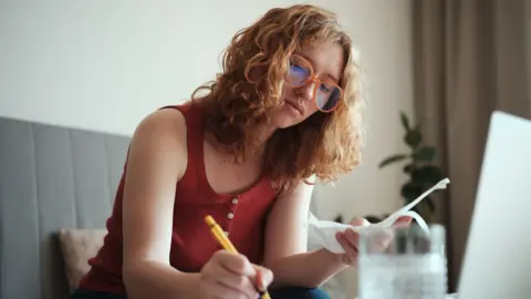 Getty Images Stock photo of a woman with glasses on and a red top writes something with a pen with bills in her other hand in front of a laptop while sitting on a sofa at home.