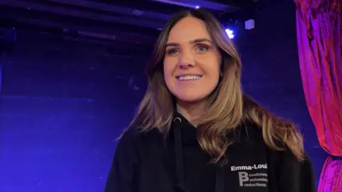 A woman with brown hair stood in front of a blue wall wearing a black hoodie. 