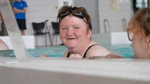 Amy is looking at the camera smiling while in the water and holding the side of the pool with both hands. Her hair is wet and she's got a pair of black goggles on her head.