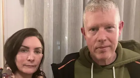 Un hombre y una mujer están uno al lado del otro frente a una cortina. La mujer, de cabello corto y oscuro, está a la izquierda. El hombre está a la derecha. Ambos llevan abrigos de invierno.