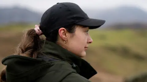 Joe Roper The side profile of Jessie Roper who is looking to the right. She is wearing a big green coat with a hood and a black cap. Her hair is tied in a plait with a pink scrunchie and she is wearing a gold hoop earring. She has a straight face and is standing outside with hills in the background which is out of focus.