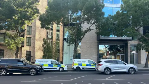 PA Media Two white Metropolitan Police Forensic Services vans parked along a street in front of a modern residential apartment building and office entrance displaying a "Tiller & Lock" sign. A police officer stands beside the rear side door of the first van.