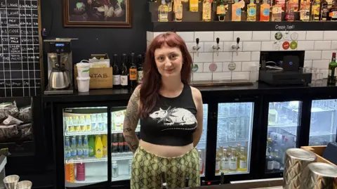 A woman with long dark red hair wears a black crop top with a white rat on the front. She is also wearing green patterned trousers while stood behind a bar with beer taps and fridges filled with glasses and bottles behind her.