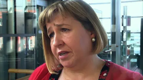 Lynne Neagle who has bobbed brown hair and is wearing a red cardigan, frowning and facing side-on to the camera