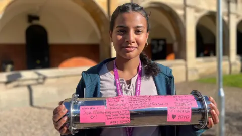 A girl with dark hair, tied back into a ponytail, wears a blue zip-up hoodie and stripe top. She smiles at the camera, standing in front of the school, while holding the time capsule. The time capsule is covered in pink coloured messages created by the students.