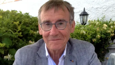Norman Esslemont Norman Esslemont smiling wearing a grey suit. He is sat in front of a green bush and white wall.