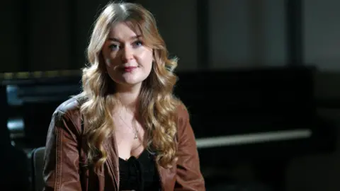 Josie Duncan has long, curly fair hair and is wearing a brown jacket over a black top. She is sitting in a darkened room looking at a camera.