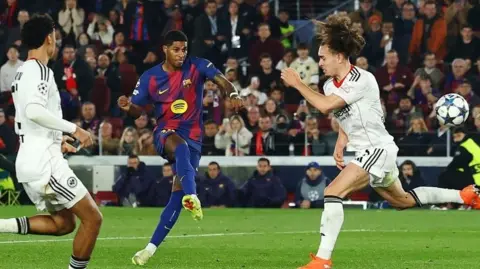 Footballer Marcus Rashford on the pitch playing for FC Barcelona. He is taking a shot as a defender to the right tries to block it and fails. Another defender to the left watches on, with a busy stadium crowd in the background
