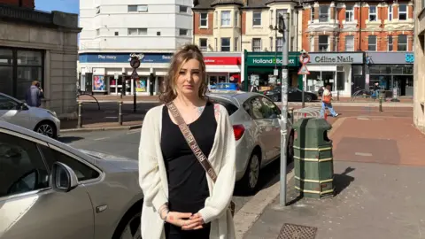 Hsin-Yi Lo A blonde woman, wearing a white cardigan and black shirt, is standing on a footpath. There is a car beside her. 