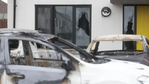 Three burnt-out cars outside a house with a yellow door, which has some broken windows