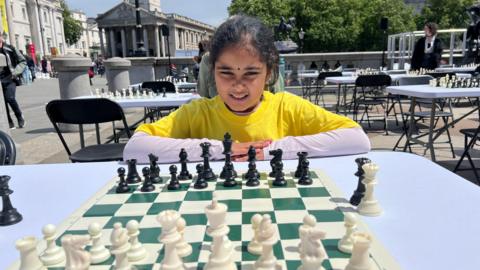 Bodhana Sivanandan is sitting down with her arms on a white table that has a chessboard on it. In the background is Trafalgar Square.