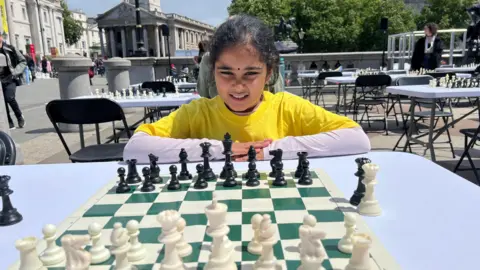 BBC Bodhana Sivanandan is sitting down with her arms on a white table that has a chessboard on it. In the background is Trafalgar Square.