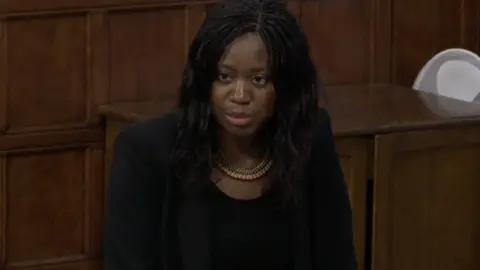 Parliament TV A woman wearing a dark suit and a gold necklace is standing in a room.
