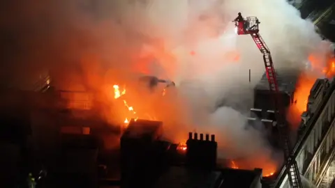 UKNIP A drone shot of someone on an aerial platform shooting water at flames on the roof of the building.