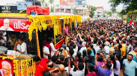 CPI (M)/X multidão de pessoas com braços esticados em direção a um ônibus vermelho decorado com guirlandas de cravo. O ônibus carrega o corpo de Achuthanandan