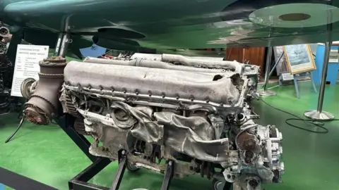 George Carden/BBC The large grey Spitfire engine which is mostly intact. There are some dents to the exhaust system and a small gash in the top cylinder where the oil leaked out. The propellers at the front also still have some of the wood from the propellers.