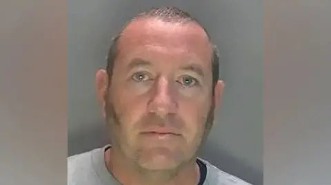 Hertfordshire Police David Carrick photographed in police custody. He is wearing a grey sweatshirt and has sideburns and a near-shaven head. 