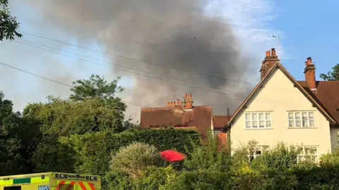AJ Fielder Black smoke from an industrial fire rising above houses and hedges in Letchworth Garden City. The roof of an ambulance can be seen on the right.