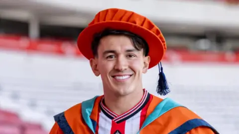 Luke O'Nien is wearing an orange graduation cap (with a dark blue tassel) and is smiling. He is wearing a Sunderland AFC red and white kit underneath his orange graduation gown and seats at the Stadium of Light are out of focus in the background. 