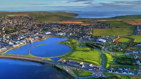 Orkney 2025 An aerial view of Orkney by the sea which shows a green landscape with homes and buildings connected by roads, large blue lakes and a running track.