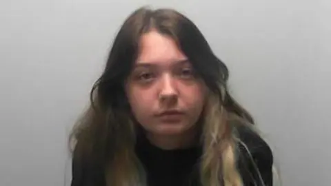 A young woman with long dark hair featuring blonde streaks sits against a plain light‑coloured wall, wearing a black sweatshirt.